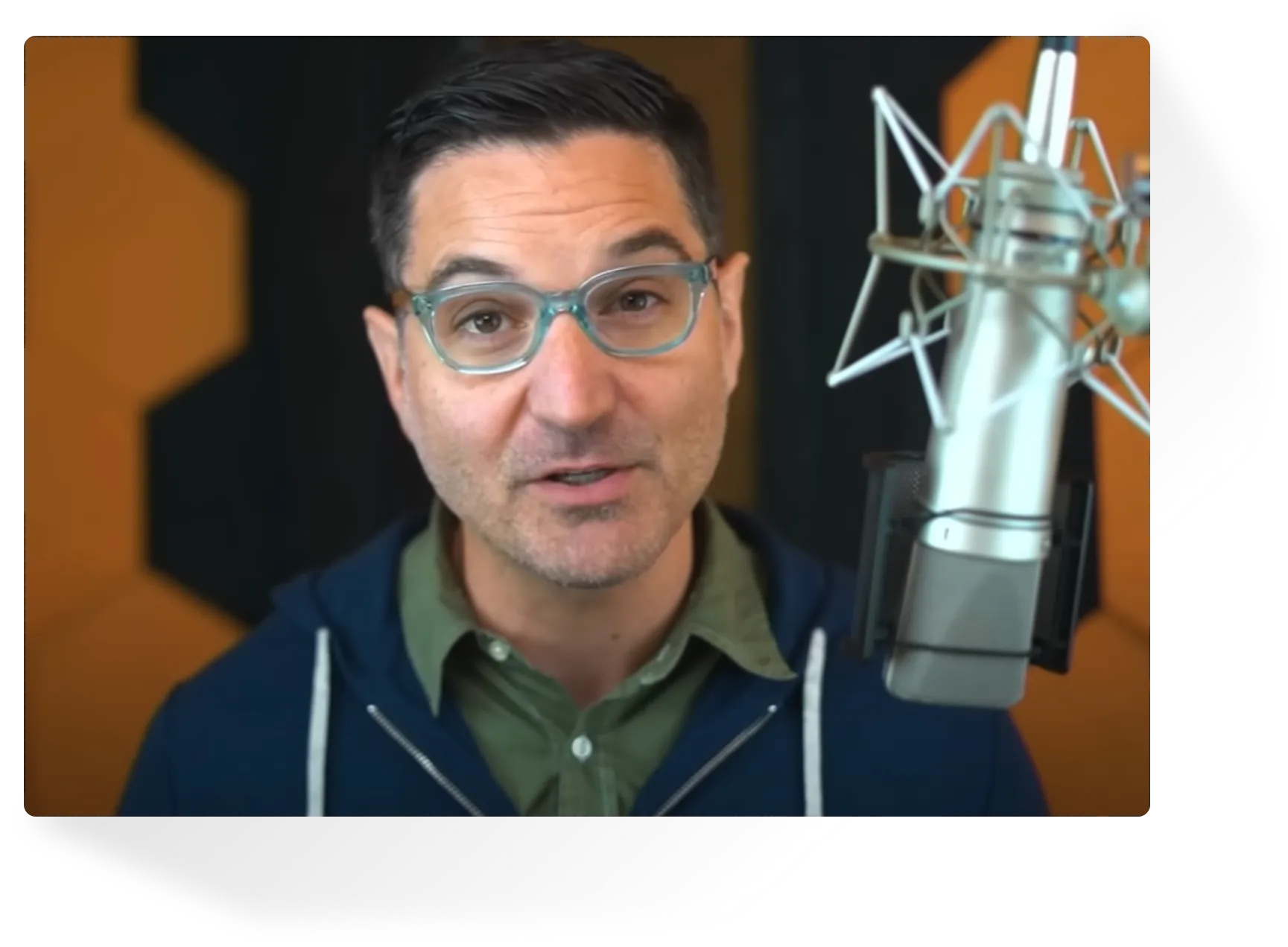 Man with eyeglasses speaking into a professional microphone with orange and black soundproofing panels in the background.